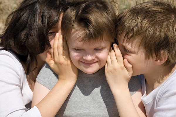Das Bild zeigt 2 Kinder die einem weiteren Kind etwas ins Ohr flüstern